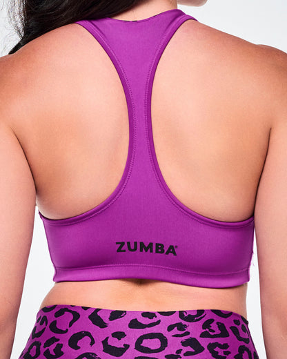 Close-up of a female model wearing a bright solid purple reversible sports bra with smaller black Zumba lettering towards the bottom hem.