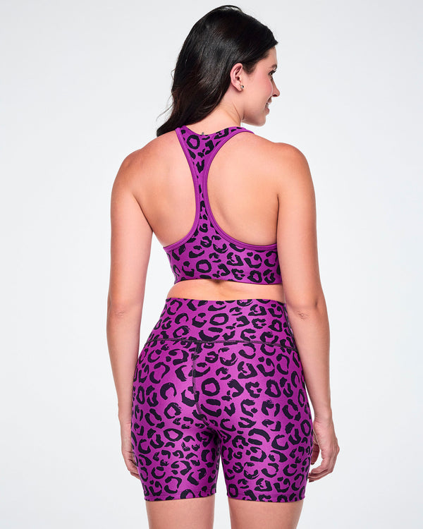 Back view of a female model wearing a matching purple and black leopard-print activewear set, featuring a reversible sports bra and high-waisted biker shorts, against a white background.