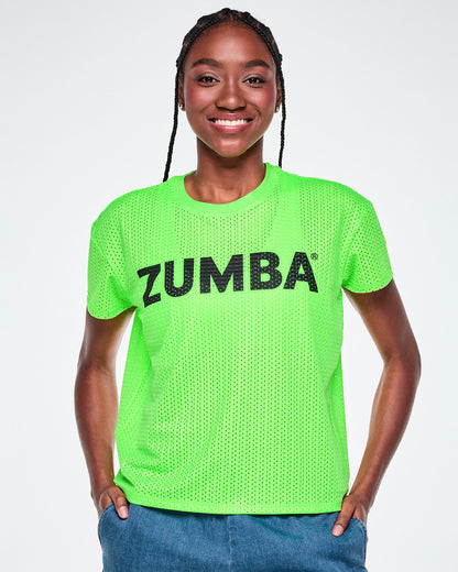 A female model wearing a bright green Zumba short-sleeve mesh top with black 'ZUMBA' text across the chest, paired with blue denim pants, against a white background.