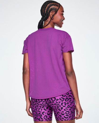 Back view of a female model wearing a purple Zumba short-sleeve mesh top, paired with purple leopard print biker shorts, against a white background.