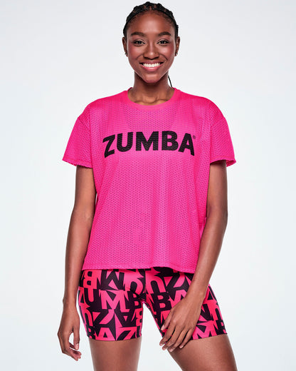 A female model wearing a bright pink Zumba short-sleeve mesh top with black 'ZUMBA' text across the chest, paired with pink and black patterned ZUMBA shorts, against a white background.