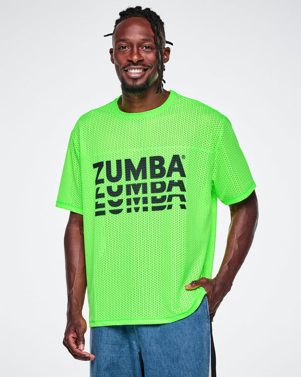 A male model wearing a neon green oversized mesh boxy top with a large black “ZUMBA” stacked logo across the chest, paired with blue denim pants, against a white background.