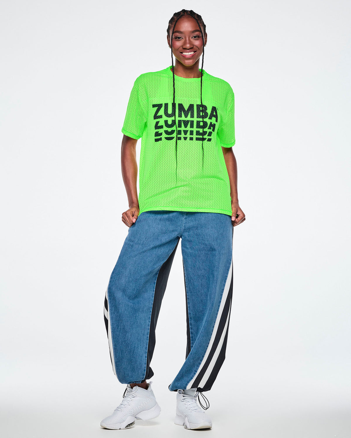 Female model in a neon green mesh tee with black 'ZUMBA' text printed three times, stacked and fading downward, paired with denim pants with black panels and white stripes.