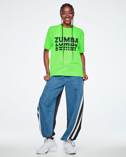 Female model in a neon green mesh tee with black 'ZUMBA' text printed three times, stacked and fading downward, paired with denim pants with black panels and white stripes.