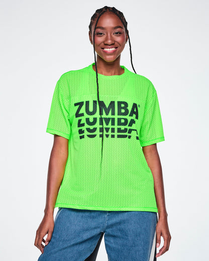 A female model wearing a neon green oversized mesh boxy top with a large black “ZUMBA” stacked logo across the chest, paired with blue denim and black pants, against a white background.