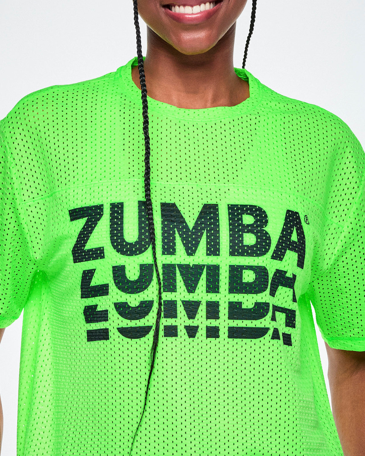 Close-up of a female model, showcasing the mesh detail of a bright green mesh short-sleeve top with a large black stacked “ZUMBA” logo across the chest.