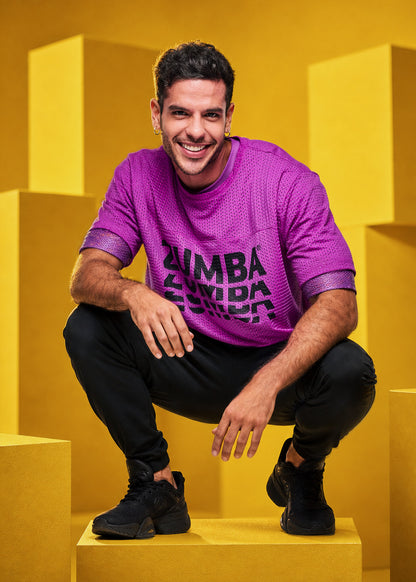A male model crouching in a studio setting with yellow geometric blocks, wearing a bright purple Zumba mesh top with black ZUMBA text, black pants, and black sneakers.