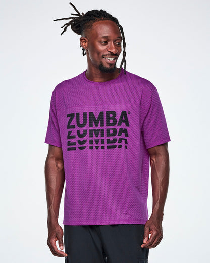 A male model in a purple oversized mesh boxy top with a large black stacked “ZUMBA” logo across the chest, paired with black pants.