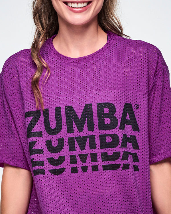 Close-up of a female model, showcasing the mesh detail of a purple mesh short-sleeve top with a large black stacked “ZUMBA” logo across the chest.