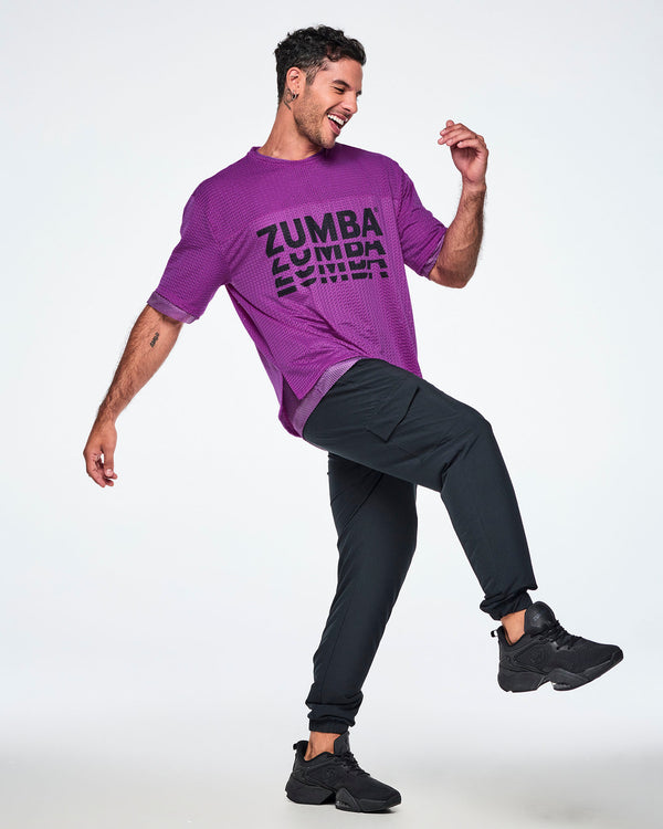 A male model wearing a purple mesh top with a black 'ZUMBA' graphic stacked on the front, paired with black pants and black sneakers on a white background.