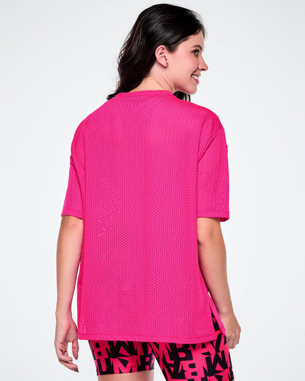 Back view of a female model wearing a hot pink oversized mesh boxy top, paired with black and pink biker shorts featuring an all-over bold “ZUMBA” lettering pattern.