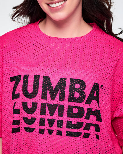 Close-up of a female model, showcasing the mesh detail of a bright pink mesh short-sleeve top with a large black stacked “ZUMBA” logo across the chest.