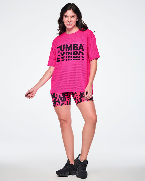 Full view of a female model wearing a hot pink oversized mesh boxy top with a large black “ZUMBA” stacked logo across the chest, paired with black and pink biker shorts featuring an all-over bold “ZUMBA” lettering pattern and black shoes.