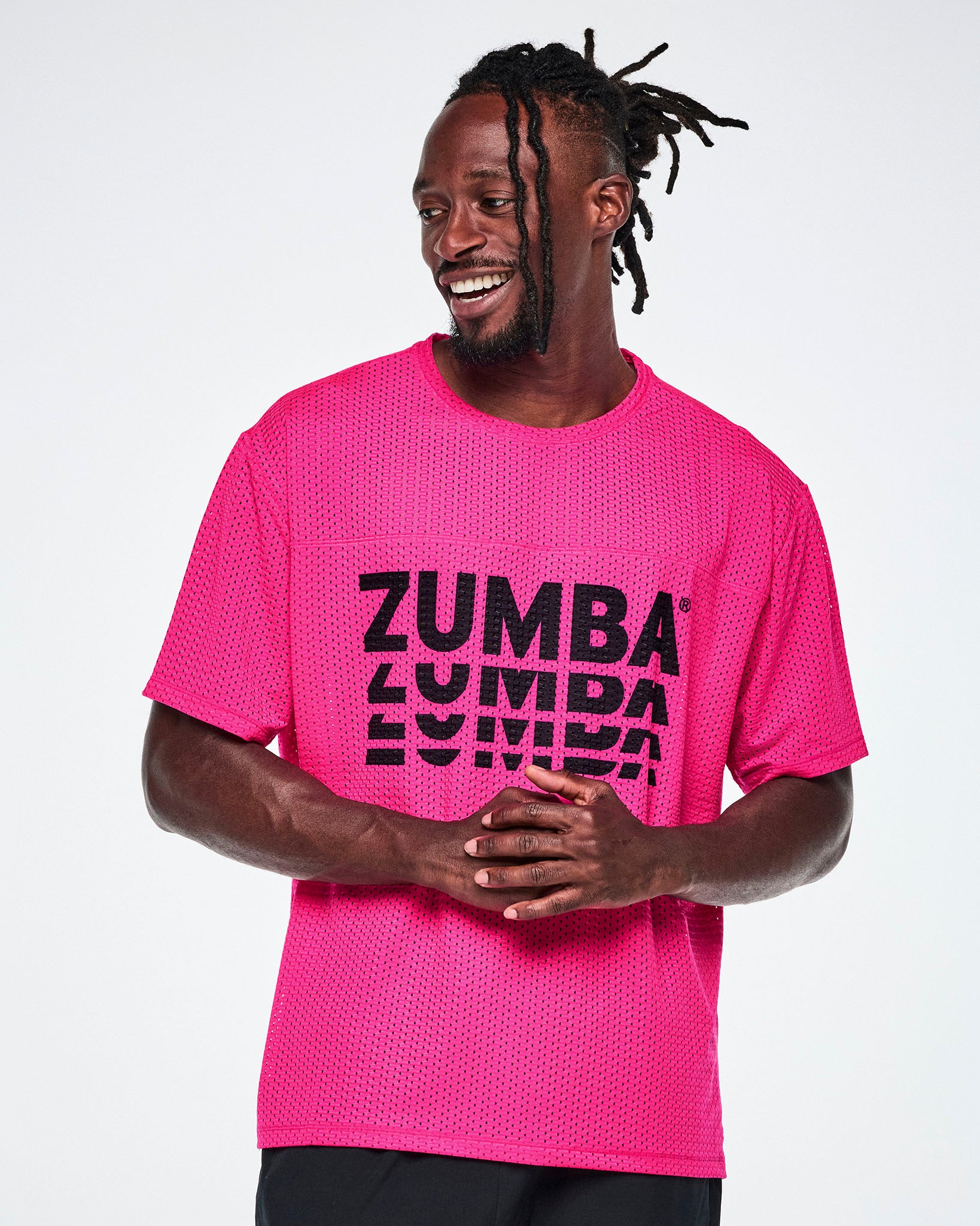 A male model wearing a neon pink oversized mesh boxy top with a large black “ZUMBA” stacked logo across the chest, paired with black pants against a white background.