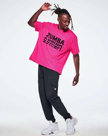 A male model wearing a bright pink mesh top with a large black stacked “ZUMBA” logo across the chest, paired with black cargo pants and white sneakers against a white background.