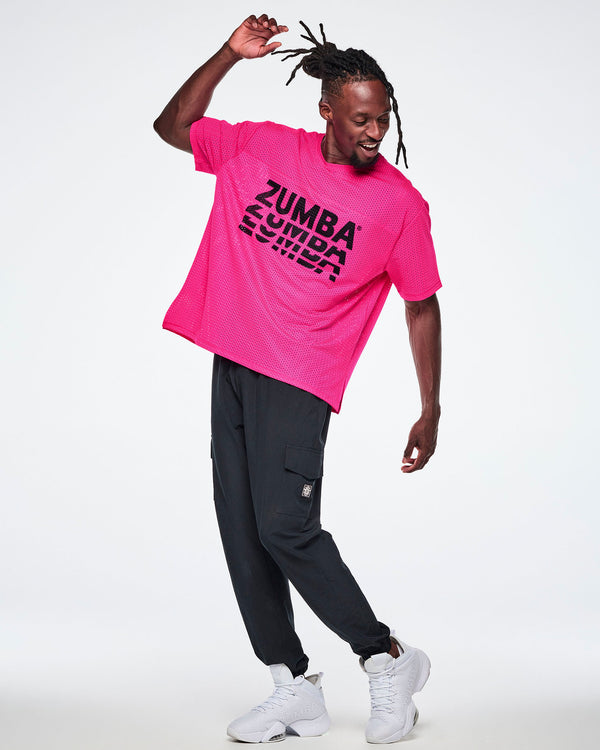 A male model wearing a bright pink mesh top with a large black stacked “ZUMBA” logo across the chest, paired with black cargo pants and white sneakers against a white background.