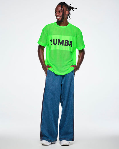 Male model with both hands in his pockets, wearing a neon green mesh tee with a solid chest band printed with black 'ZUMBA' text, paired with wide-leg denim pants with dark side panels.