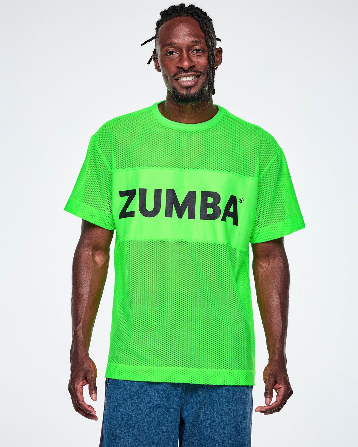A male model wearing a bright green Zumba mesh top with a wide band across the chest featuring the black ZUMBA text, paired with blue denim pants.