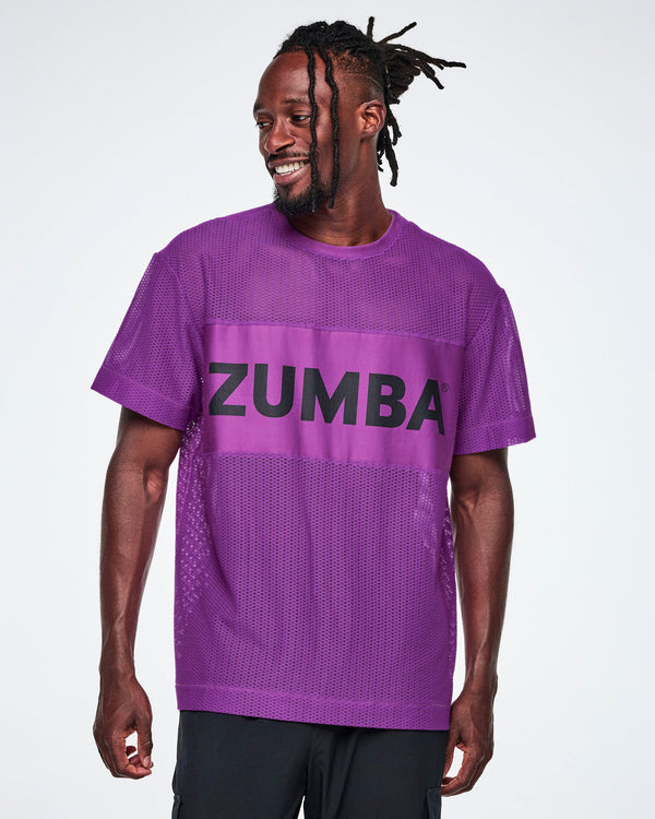 A male model wearing a purple Zumba mesh top with a wide band across the chest featuring the black ZUMBA text, paired with black pants.