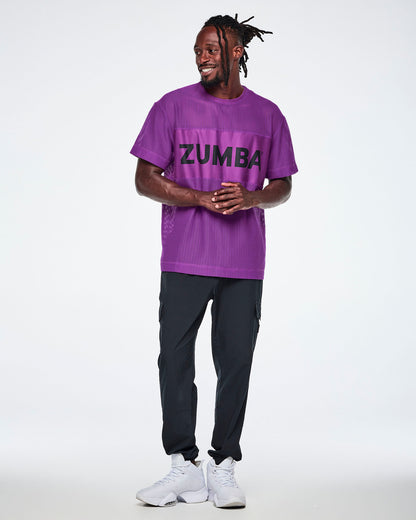 Full view of a male model wearing a purple Zumba mesh top with a wide band across the chest featuring the black ZUMBA text, paired with black pants and white shoes.