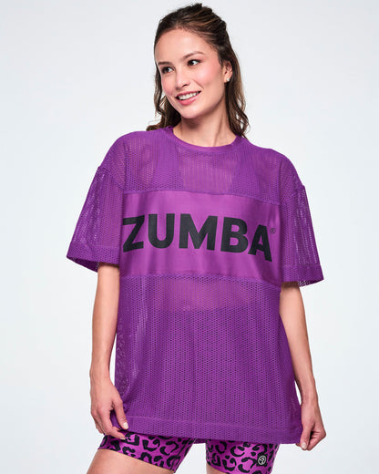 A female model wearing a purple Zumba mesh top with a wide band across the chest featuring the black ZUMBA text, paired with black and purple leopard print biker shorts.