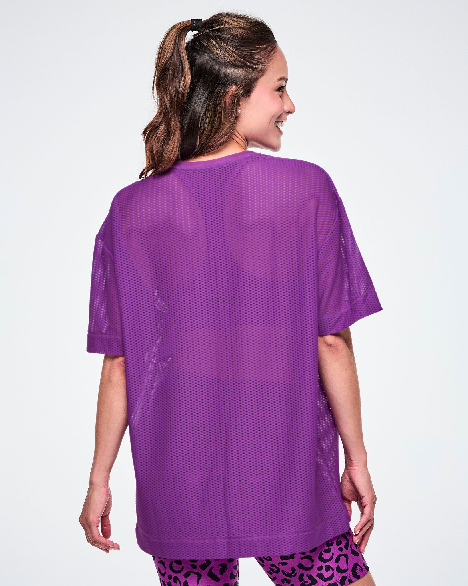 Back view of a female model wearing a purple Zumba mesh top, paired with black and purple leopard print biker shorts.