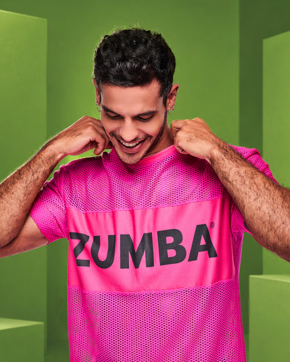 Male model with hands on his collar, wearing a bright pink Zumba mesh top featuring a wide chest band with black “ZUMBA” text, posed against a green geometric studio background.