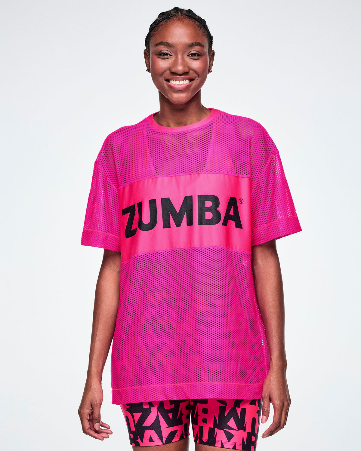 A female model wearing a bright pink Zumba mesh top with a wide band across the chest featuring the black ZUMBA text, paired with black and pink ZUMBA patterned biker shorts.