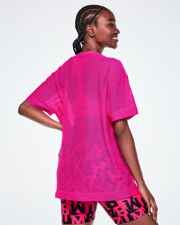 Back view of a female model wearing a bright pink Zumba mesh top, paired with black and pink ZUMBA patterned biker shorts.