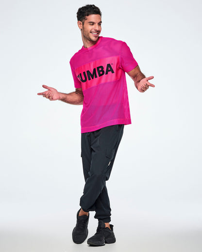 Full view of a male model wearing a bright pink Zumba mesh top with a wide band across the chest featuring the black ZUMBA text, paired with black pants and black shoes.