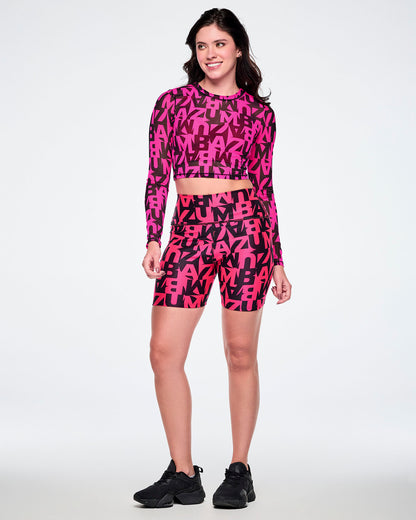 Full view of a female model wearing a matching two-piece activewear set featuring a long-sleeve mesh crop top and high-waisted biker shorts, both printed with a bold abstract pink and black ZUMBA lettering and paired with black shoes.