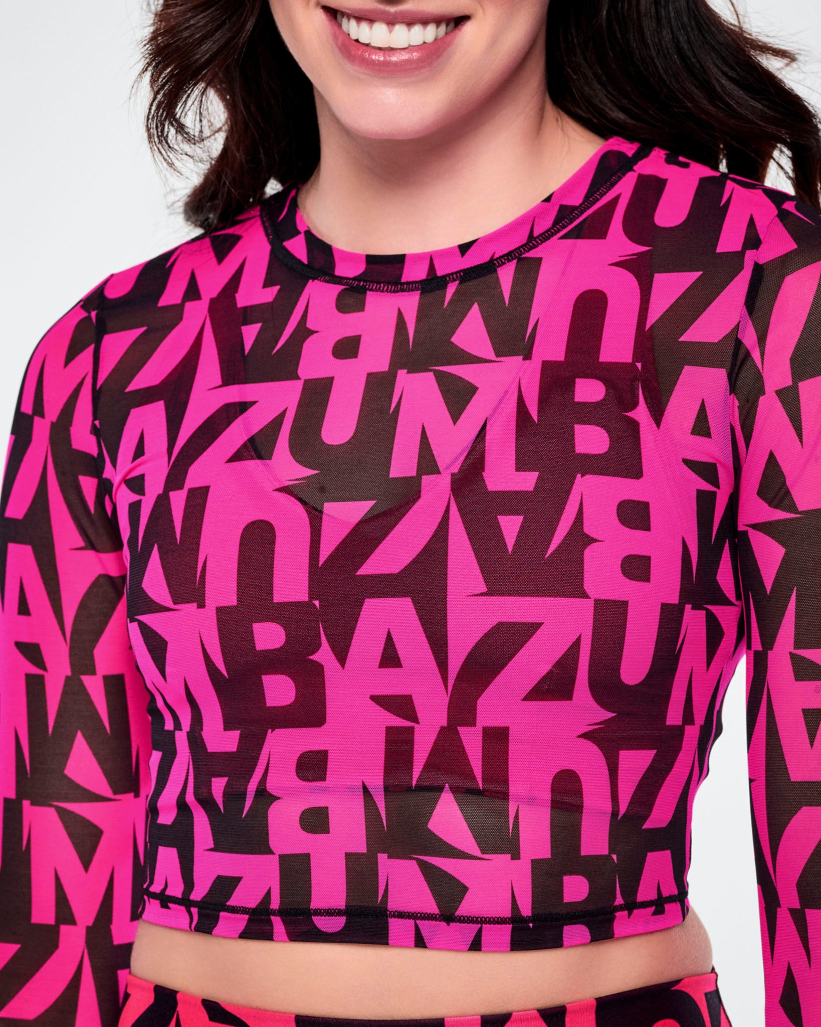 Close-up of a female model wearing a long-sleeve sheer mesh crop top with an all-over abstract pink and black ZUMBA pattern.