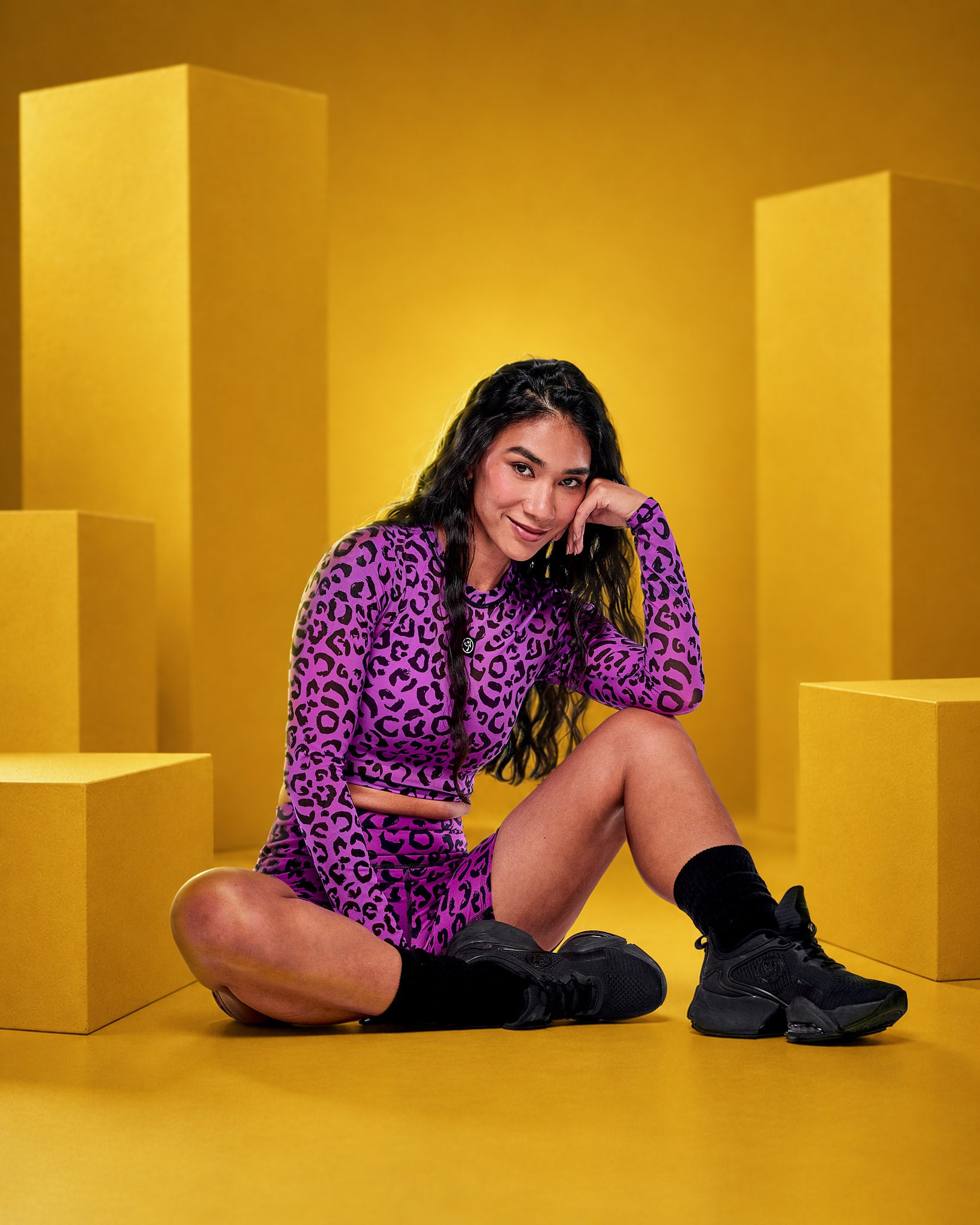 A female model sits cross-legged on a yellow studio floor surrounded by yellow geometric blocks, wearing a purple and black leopard-print long-sleeve mesh crop top and matching biker shorts with black socks and black sneakers.