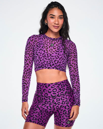 A female model wearing a matching purple and black leopard-print activewear set, featuring a long-sleeve mesh crop top and high-waisted biker shorts, against a white background.
