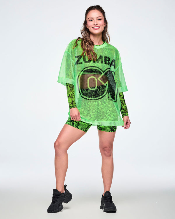 Female model in a neon green oversized mesh V-neck top with black 'ZUMBA' text over a large outlined '01' graphic, layered over a green snakeskin-print long-sleeve top and matching biker shorts, with black shoes.