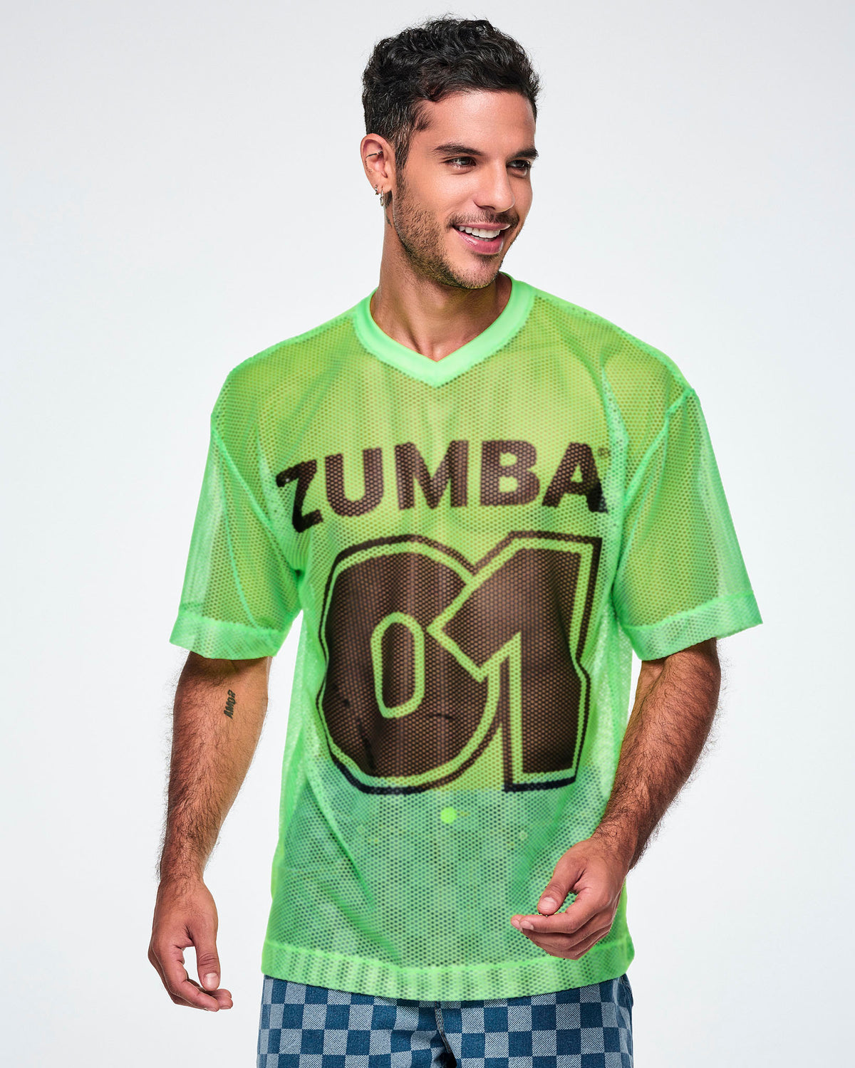 A male model wearing a green mesh Zumba tee with a large black “ZUMBA 01” on the front, paired with blue checkered denim pants.