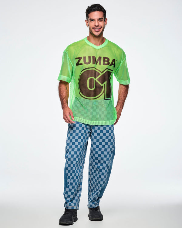 Full view of a male model wearing a green mesh Zumba tee with a large black “ZUMBA 01” on the front, paired with blue checkered denim pants and black shoes.