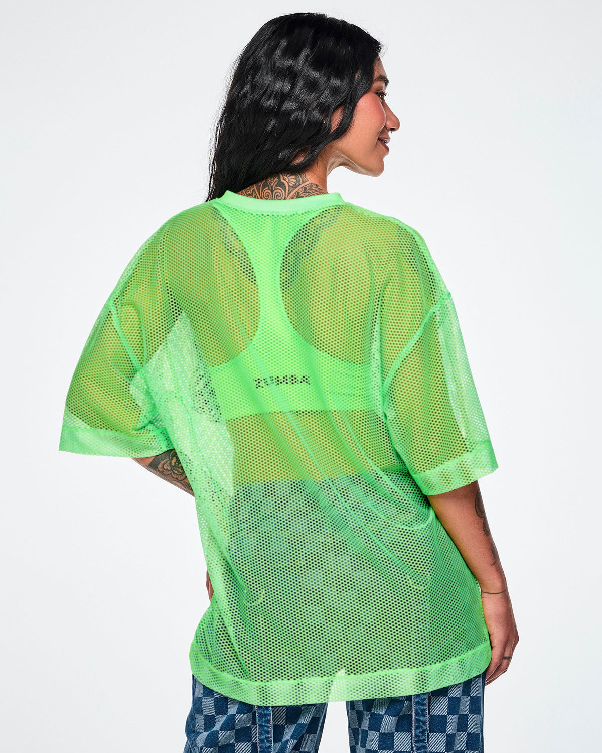 Back view of a female model wearing a green mesh Zumba top with a large black "ZUMBA 01" printed on the front, paired with blue checkered denim pants.