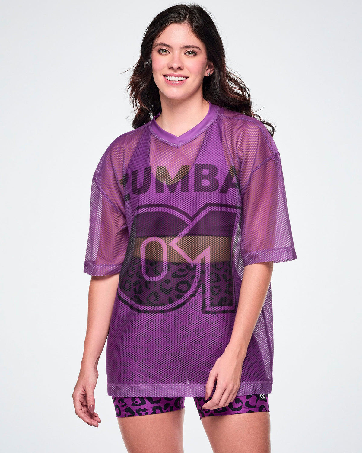 A female model wearing a purple mesh Zumba top with a large black "ZUMBA 01" printed on the front, paired with purple leopard print high-waisted biker shorts.