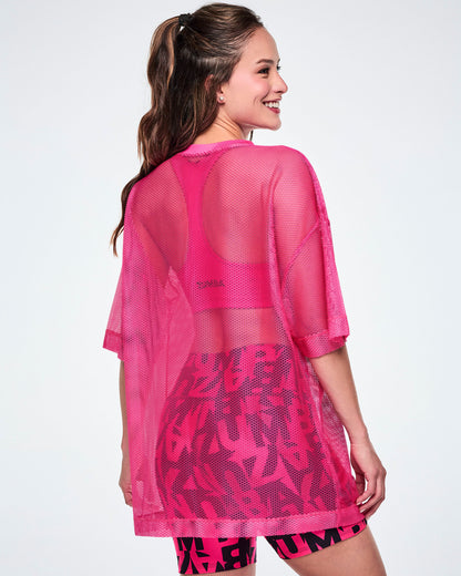 Back view of female model wearing a pink mesh Zumba top, paired with black-and-pink high-waisted biker shorts featuring an allover bold “ZUMBA” lettering pattern.