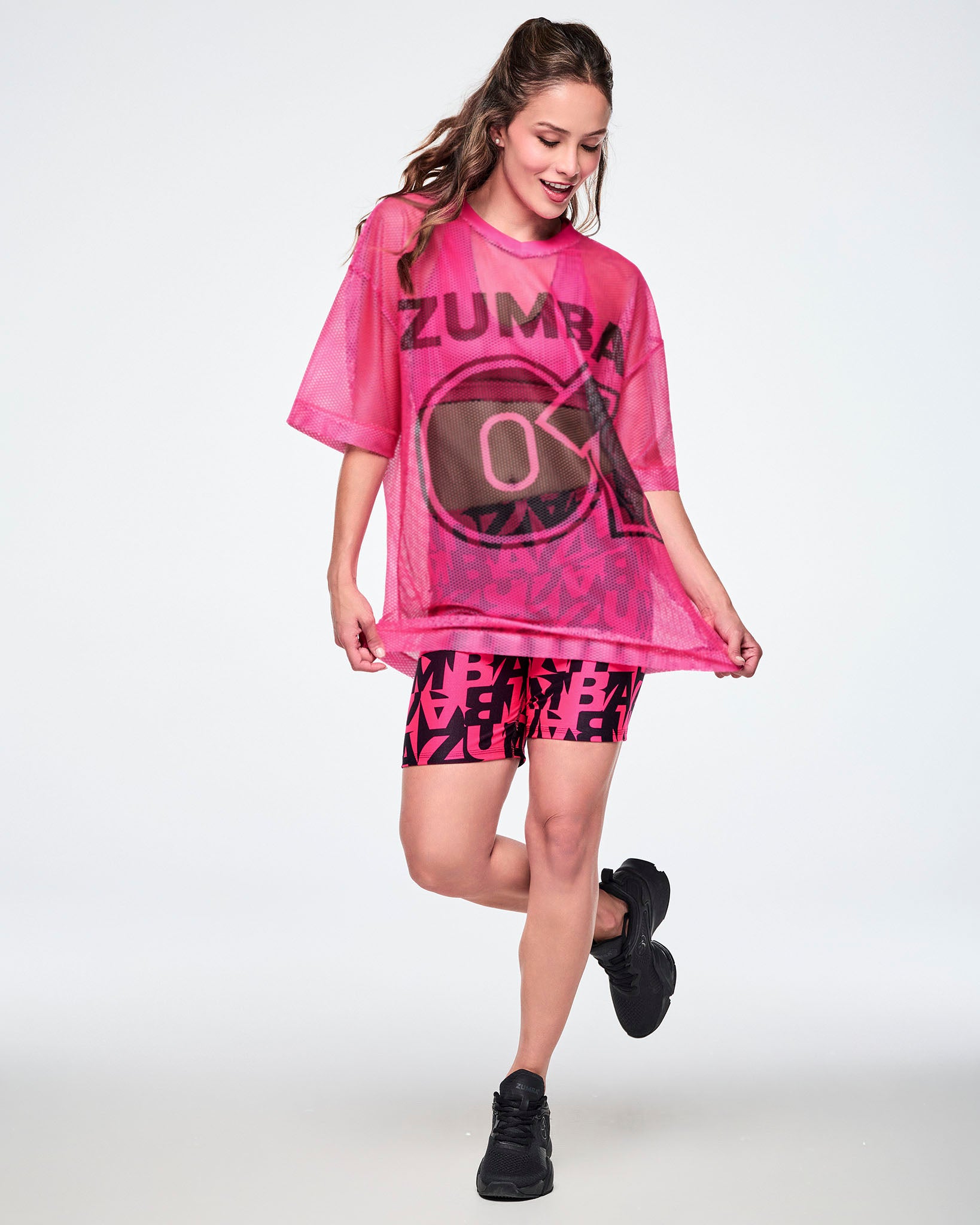 Full view of a female model wearing a pink mesh Zumba top with a large black “ZUMBA 01” on the front, paired with black-and-pink high-waisted biker shorts featuring an all-over bold “ZUMBA” lettering pattern, and black shoes.