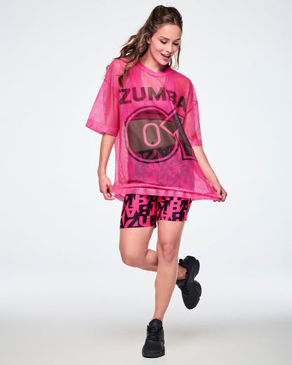 Full view of a female model wearing a pink mesh Zumba top with a large black “ZUMBA 01” on the front, paired with black-and-pink high-waisted biker shorts featuring an all-over bold “ZUMBA” lettering pattern, and black shoes.