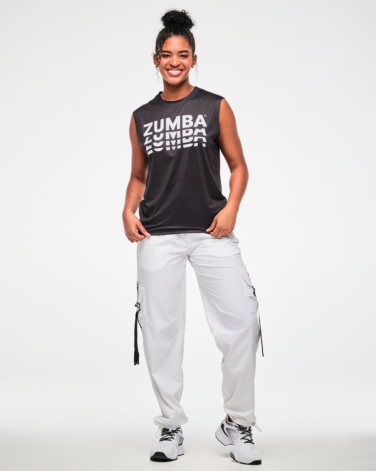 Black sleeveless muscle tee with a repeating white 'ZUMBA' logo, worn by a female model with white cargo pants and white shoes.