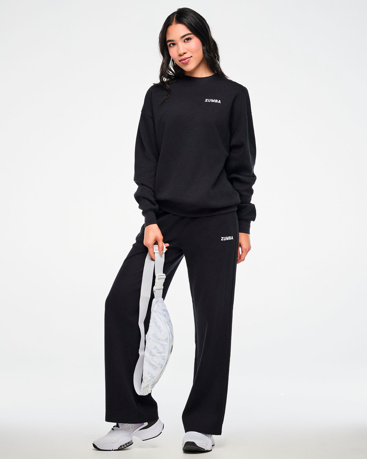 Black waffle-knit crewneck sweatshirt and matching sweatpants with a small white 'ZUMBA' logo, worn by a female model carrying a white 'ZUMBA'-print crossbody bag.