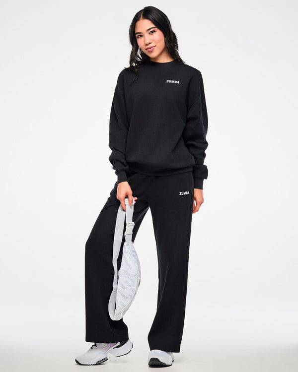 Black waffle-knit crewneck sweatshirt and matching sweatpants with a small white 'ZUMBA' logo, worn by a female model carrying a white 'ZUMBA'-print crossbody bag.