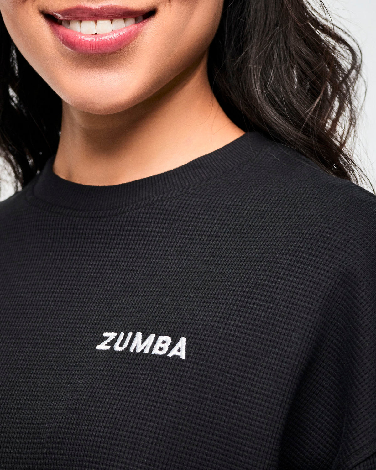 Close-up of the small white 'ZUMBA' logo on the black waffle-knit crewneck sweatshirt, female model.