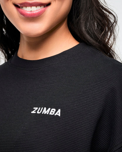 Close-up of the small white 'ZUMBA' logo on the black waffle-knit crewneck sweatshirt, female model.