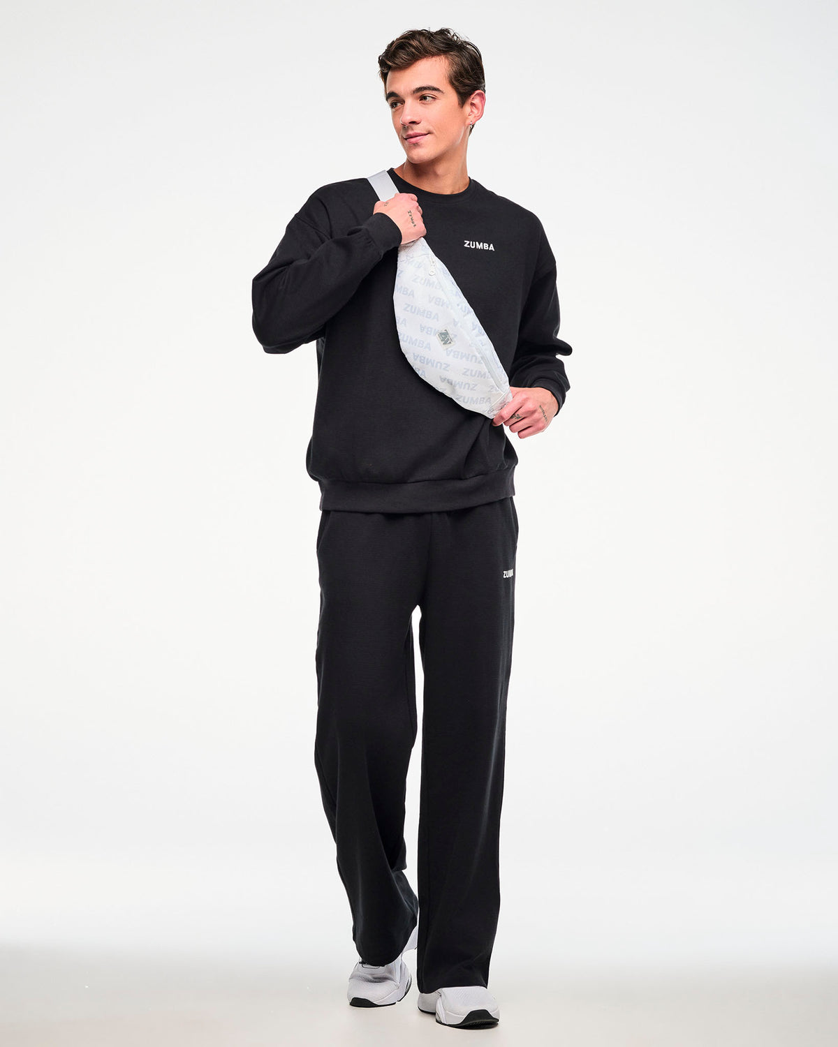 Male model in a black waffle-knit crewneck sweatshirt and matching sweatpants with a small white 'ZUMBA' logo, carrying a white 'ZUMBA'-print crossbody bag.
