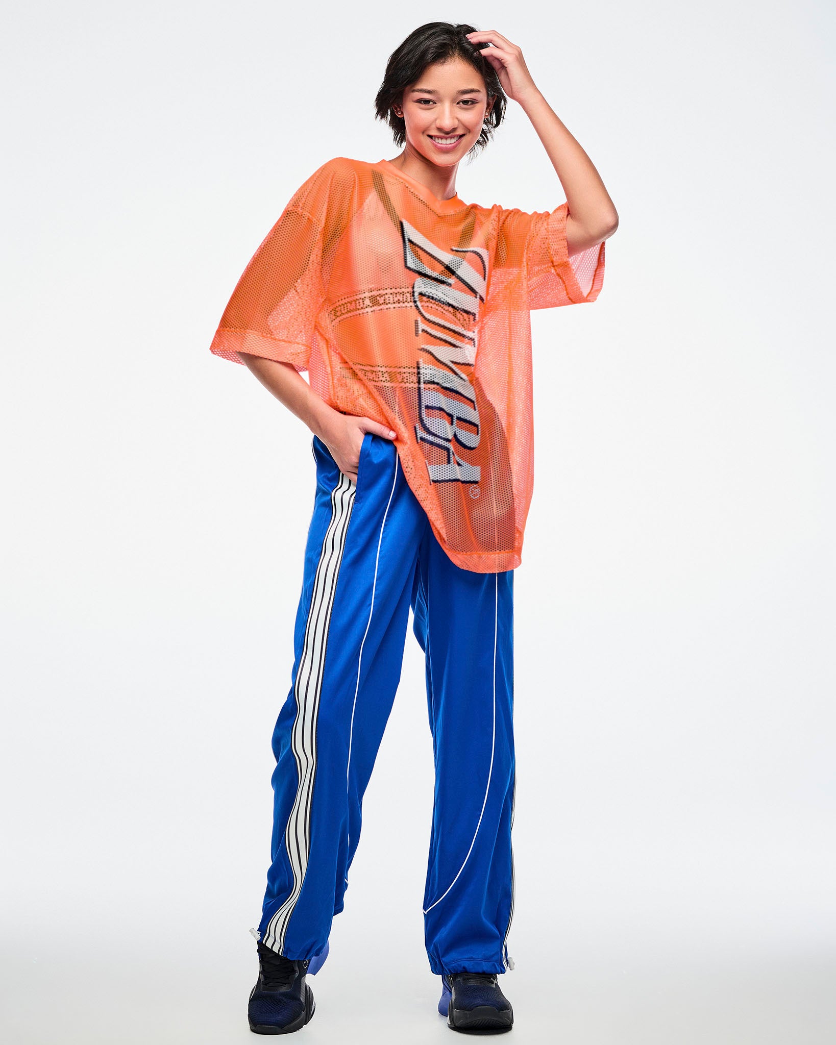 Full view of a female model wearing an oversized orange mesh Zumba top with bold vertical “ZUMBA” silver lettering paired with blue and white striped pants on a white background. 