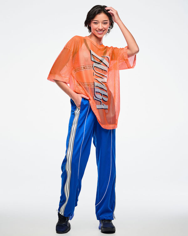 Full view of a female model wearing an oversized orange mesh Zumba top with bold vertical “ZUMBA” silver lettering paired with blue and white striped pants on a white background. 